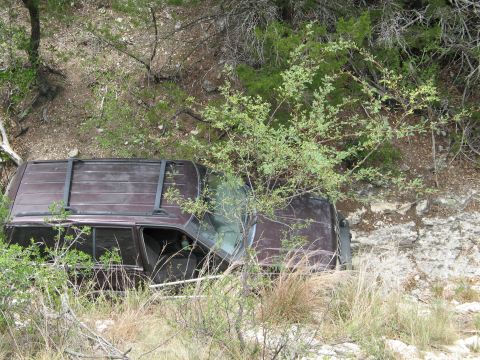 1995 Ford Bronco - A maroon SUV is seen crashed off a dirt road, surrounded by dense vegetation and trees.