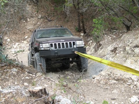 1995 Ford Bronco - A dark Jeep Grand Cherokee navigates a challenging rocky trail, with a yellow tow strap connected to its front bumper.