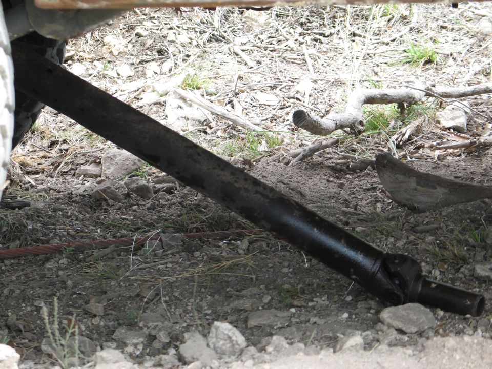 Driveshaft visible under a vehicle, resting on the ground with rocks and debris