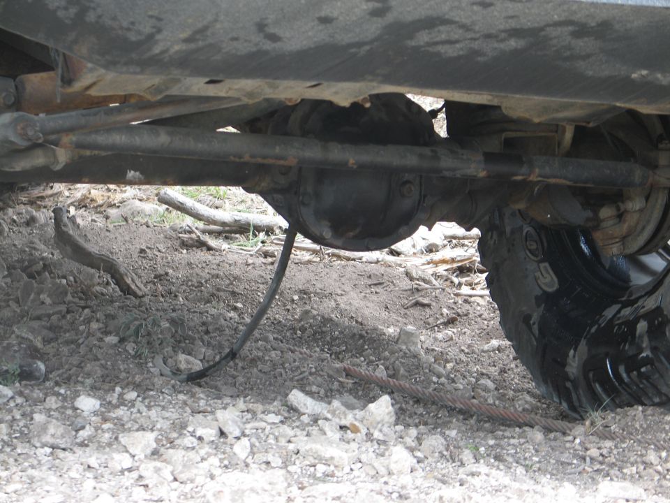 Undercarriage of a vehicle showing the rear differential, suspension components, and a large tire. A cable lies on the ground