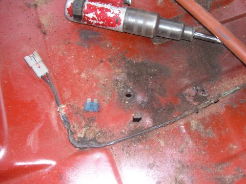 1985 Ford F-150 - Close-up of a vehicle's red floor shows rust, a fuse, and wiring, with a tool in the background.