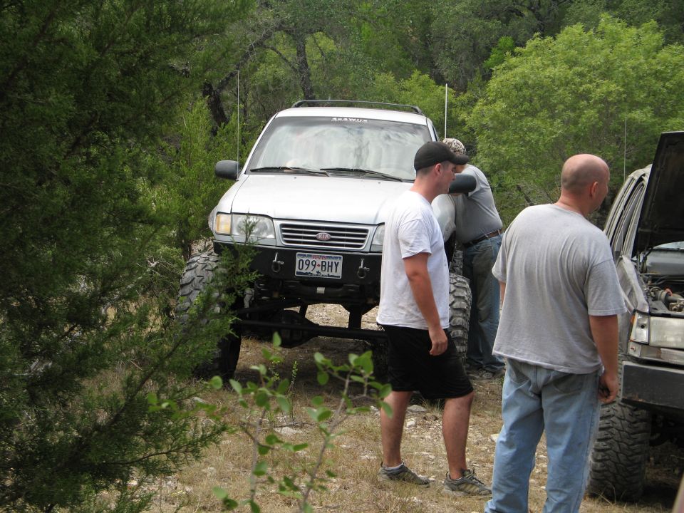 Modified SUV with large tires parked off-road; two men stand near it, another works on an open-hood vehicle