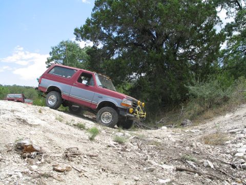 1995 Ford Bronco - A red and grey Ford Bronco navigates a rocky incline on a dirt trail, with another vehicle visible in the background.