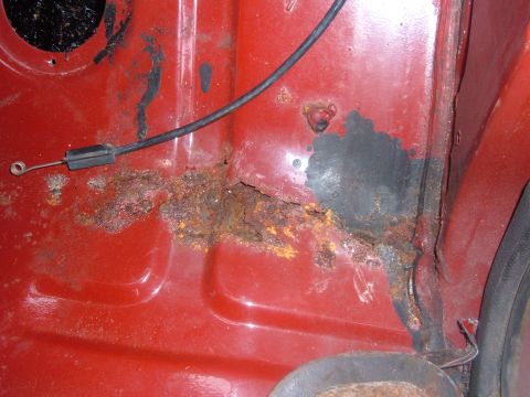 1985 Ford F-150 - Close-up view of rusted metal and a cable on a red vehicle part, showing extensive corrosion.