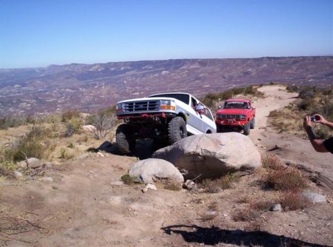1995 Ford Bronco - Two off-road vehicles, a white Bronco and a red pickup, traverse a dirt path in a , mountainous area.