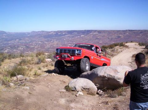 1995 Ford Bronco - A red off-road truck maneuvers over rocks on a dusty trail with a mountainous landscape in the background.