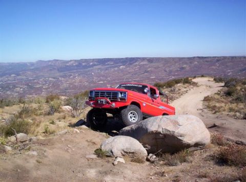 1995 Ford Bronco - A red off-road truck navigates a rocky trail with a scenic mountain vista in the background. The vehicle appears to be from the early 1980s.
