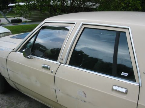1985 Ford LTD Crown Victoria - Side view of a 1985 Ford Crown Victoria LTD sedan, showing its doors and windows. The car is tan and has visible bodywork.