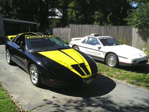 1984 Pontiac Fiero - Two Pontiacs, a black and yellow Trans Am and a white Fiero, are parked side-by-side on a driveway.