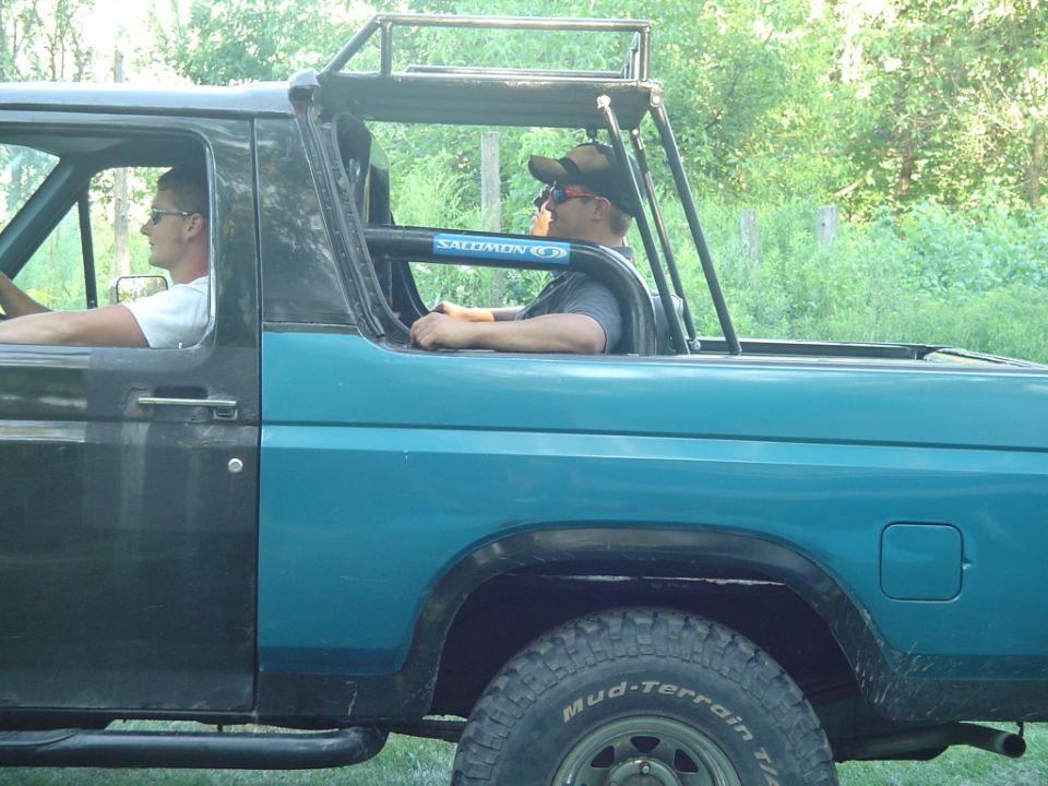 Side view of teal topless Ford Bronco with roll bar and two occupants on grass