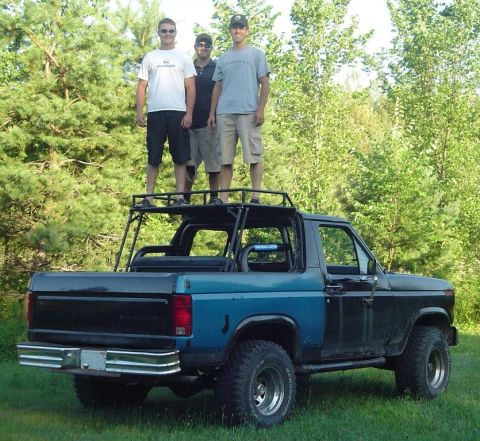 1986 Ford Bronco - Three men stand on the roof rack of a modified Ford Bronco parked outdoors.