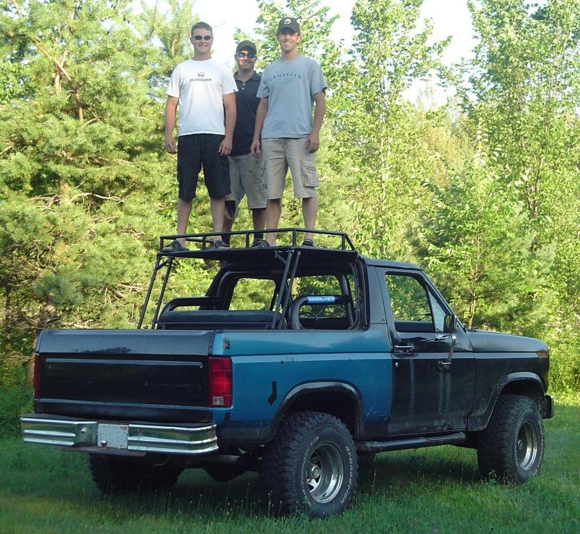 Blue and black topless Ford Bronco with three men standing on roof rack, parked on grass