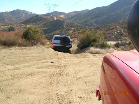 1985 Ford Bronco - A white SUV navigates a sandy trail in a mountainous, arid landscape, with a red vehicle partially visible in the foreground.