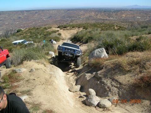 1985 Ford Bronco - A white off-road truck navigates a challenging rocky trail with a scenic overlook in the background.