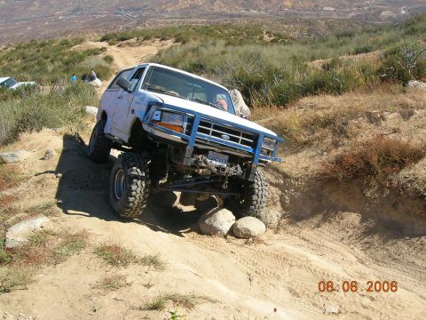 1985 Ford Bronco - White off-road SUV navigates a challenging rocky incline on a sunny day.
