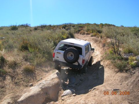 1985 Ford Bronco - A white 1985 Ford Bronco navigates a challenging off-road trail with a spare tire mounted on the back.