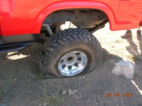 1985 Ford Bronco - A flat tire is visible on the rear wheel of a red truck, resting on dirt ground.