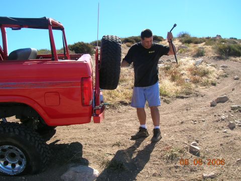 1985 Ford Bronco - A man stands next to a red 1985 Ford Bronco, holding a jack, with a spare tire mounted on the rear.
