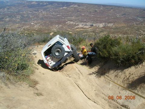 1985 Ford Bronco - A white SUV is overturned on a steep dirt incline, with two individuals observing the scene.