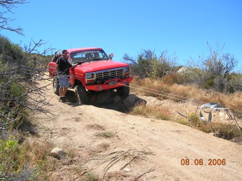 1985 Ford Bronco - A red pickup truck is being winched up a rocky, off-road trail under a clear blue sky.