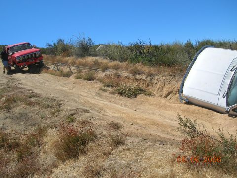 1985 Ford Bronco - A red pickup truck is seen towing a white SUV that has become stuck on a dirt embankment under a clear blue sky.