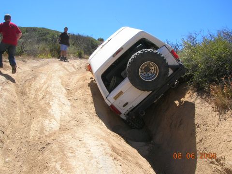 1985 Ford Bronco - A white SUV is stuck on its side in a deep dirt rut with two onlookers observing the scene.