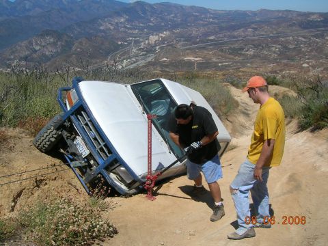 1985 Ford Bronco - Two men attempt to right a white Ford Bronco that has rolled onto its side on a steep, dusty trail.