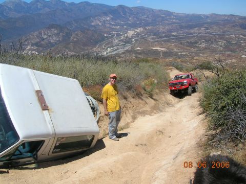 1985 Ford Bronco - A white SUV is on its side on a steep dirt road, with a red truck navigating the trail behind it.