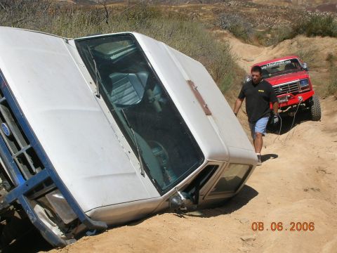 1985 Ford Bronco - A white SUV has rolled onto its side on a dirt trail, with a red Ford Bronco and a man standing nearby.