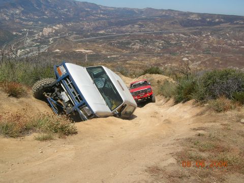 1985 Ford Bronco - Two off-road vehicles navigate a steep, dusty trail with a dramatic mountain vista.