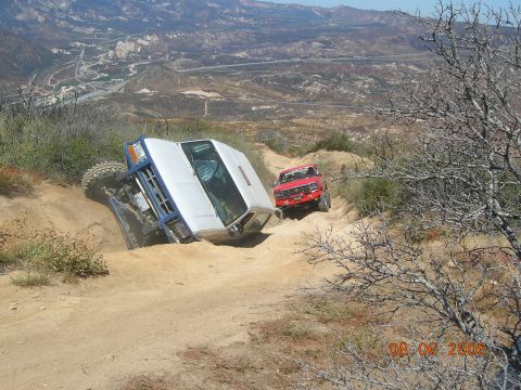 1985 Ford Bronco - Two off-road vehicles navigate a challenging dirt trail with steep inclines and a dramatic drop-off.