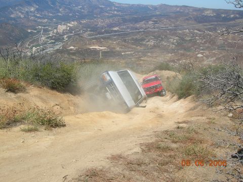 1985 Ford Bronco - Two off-road vehicles navigate a steep, dusty trail with a valley and highway visible in the background.