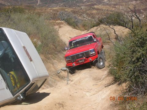 1985 Ford Bronco - Red pickup truck tows white vehicle out of a ditch on a dirt trail.