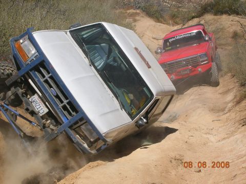 1985 Ford Bronco - A white Ford Bronco is overturned on a dirt trail, with a red Ford Bronco following closely behind.
