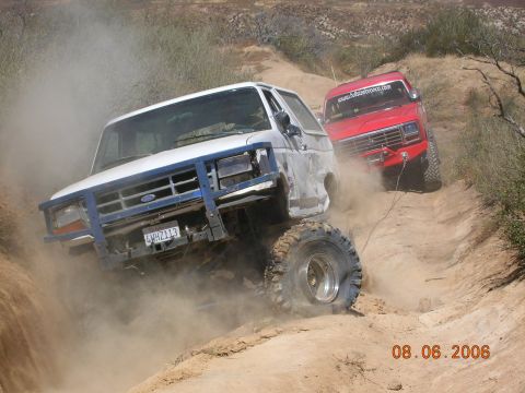 1985 Ford Bronco - Two Ford Broncos navigate a challenging off-road trail, kicking up dust as they climb.