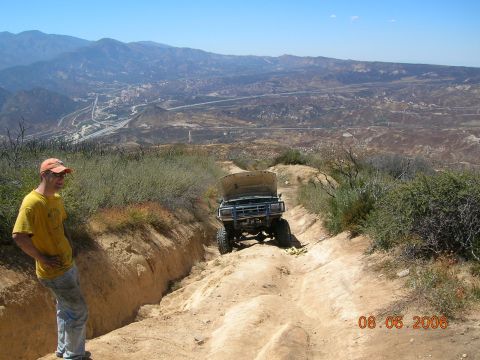 1985 Ford Bronco - A man stands by an off-road vehicle navigating a challenging dirt path with a scenic mountainous vista.