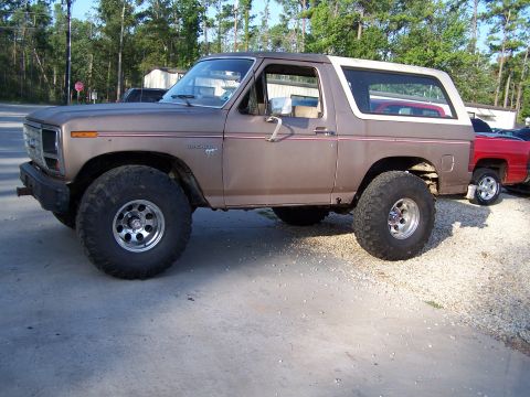 1981 Ford Bronco - This lifted Ford Bronco is parked next to a red pickup truck on a gravel and concrete surface.