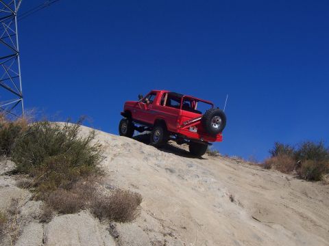 1985 Ford Bronco - A red 1985 Ford Bronco navigates an off-road trail, ascending a steep, rocky incline under a clear blue sky.