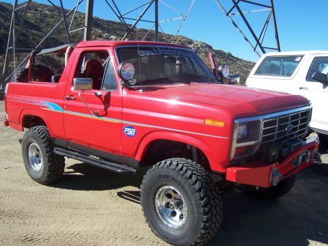 1985 Ford Bronco - A lifted red Ford Bronco is parked outdoors next to a white vehicle under a large metal structure.