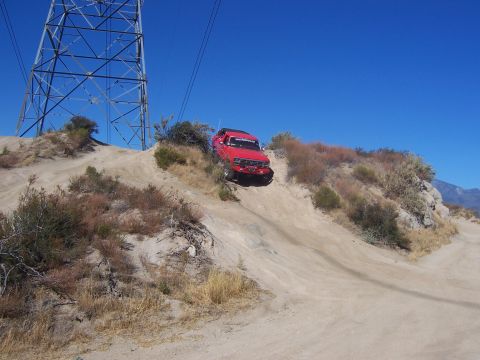 1985 Ford Bronco - A red SUV navigates a steep dirt path under a clear blue sky, with a large power pylon nearby.