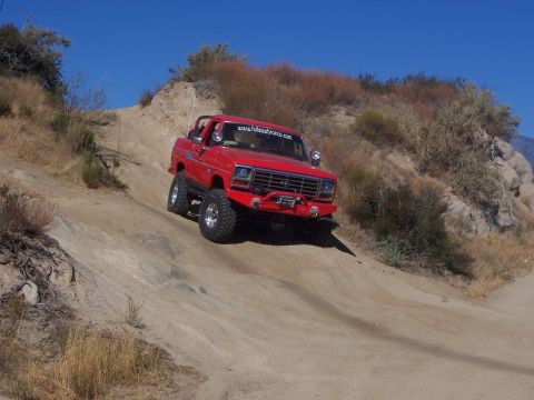 1985 Ford Bronco - A red SUV navigates a steep, dusty trail under a clear blue sky.