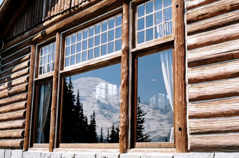 1979 Ford Bronco - A log cabin's windows reflect a mountain landscape and pine trees under a blue sky.