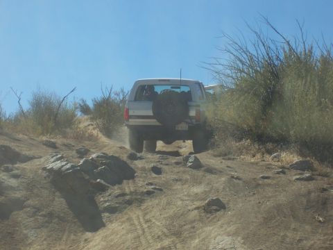 1985 Ford Bronco - A silver 1985 Ford Bronco navigates a dusty, off-road trail with dense brush on either side.