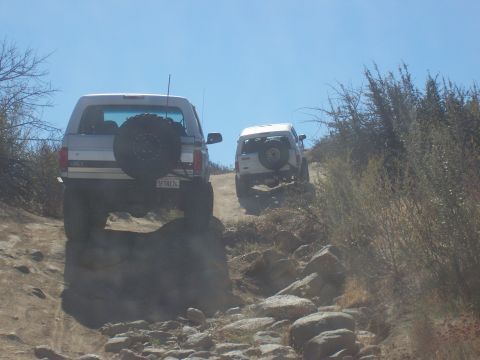 1985 Ford Bronco - Two SUVs navigate a challenging off-road trail under a clear blue sky.