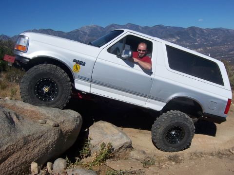 1985 Ford Bronco - A man drives a white Ford Bronco over large rocks on a sunny day with mountains in the background.