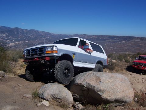 1985 Ford Bronco - White Ford Bronco climbs over large rocks on a trail, with a red Ford truck behind it.