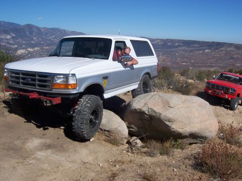 1985 Ford Bronco - A white Ford Bronco navigates a rocky trail with a red truck in the background.