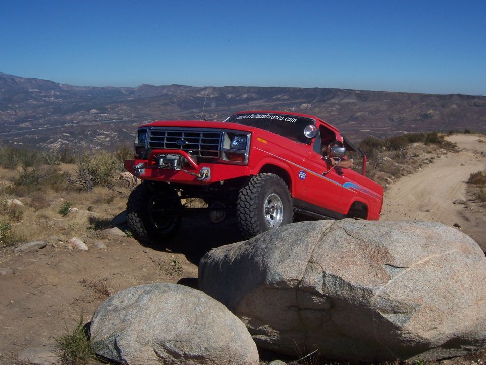 Red Ford Bronco with custom bumper and winch climbing over large rocks on a dirt trail