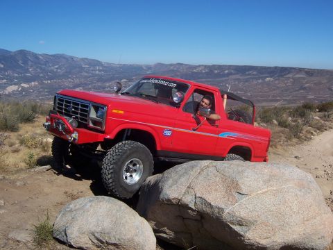1985 Ford Bronco - A red Ford Bronco is shown off-road, climbing over large rocks under a clear blue sky.
