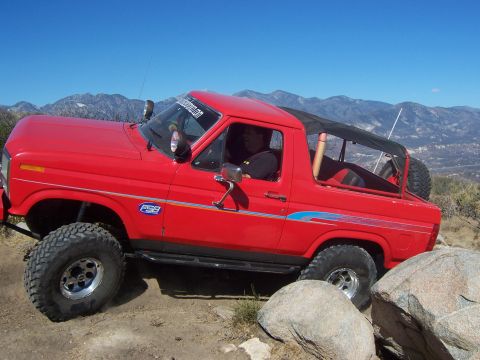 1985 Ford Bronco - This red 1985 Ford Bronco is navigating a challenging rocky terrain under a clear blue sky.